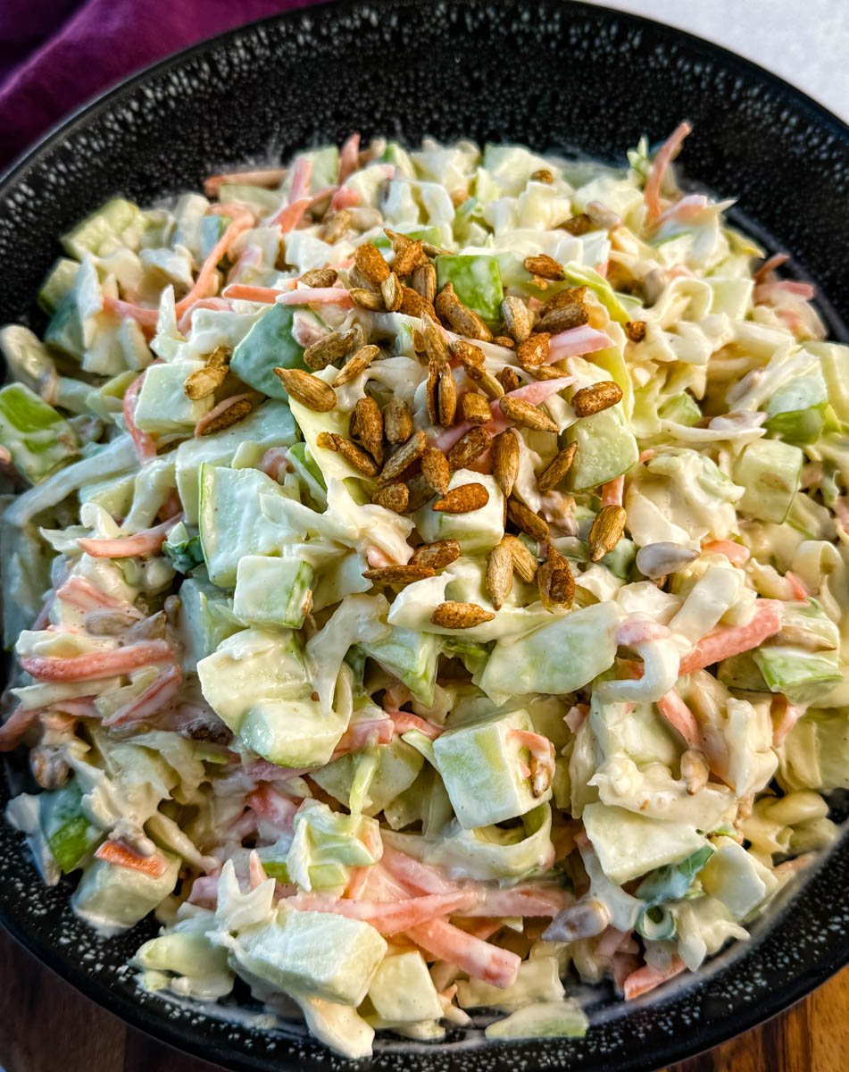 Close-up of creamy apple coleslaw topped with toasted sunflower seeds, showing chopped green apples, shredded cabbage, and carrots in a creamy dressing.