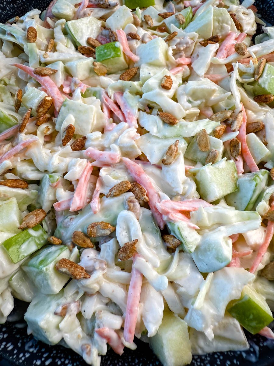 Close-up of creamy apple coleslaw topped with toasted sunflower seeds, showing chopped green apples, shredded cabbage, and carrots in a creamy dressing.