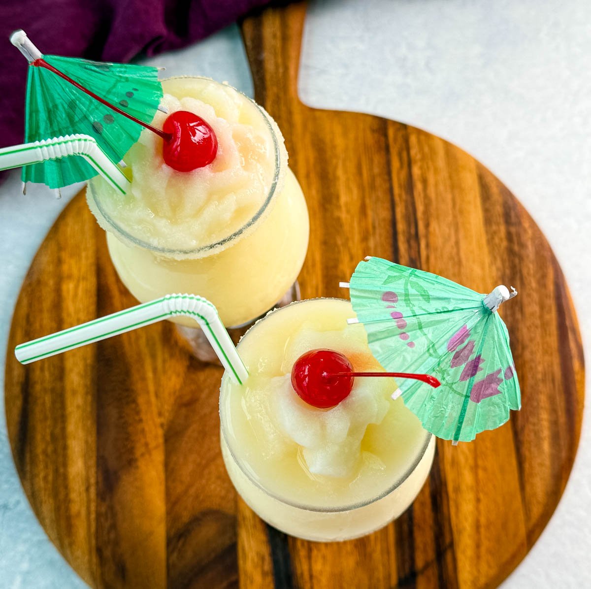 Two frozen piña coladas served in glasses on a wooden board, topped with cherries and paper umbrellas.