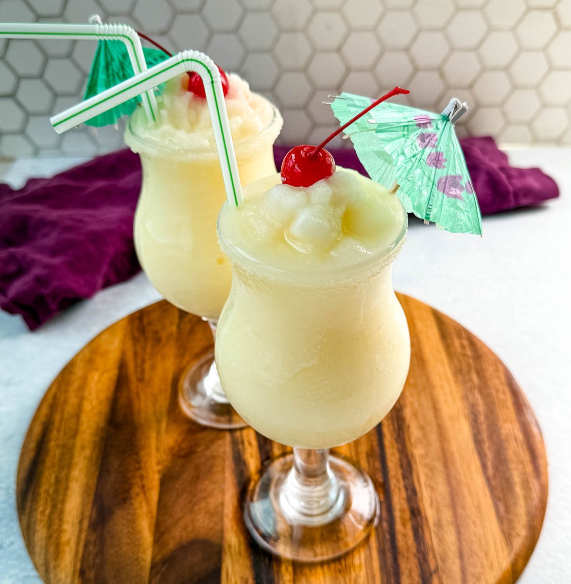 Close-up of creamy piña coladas in curved glasses with straws, cherries, and umbrellas, ready to serve.