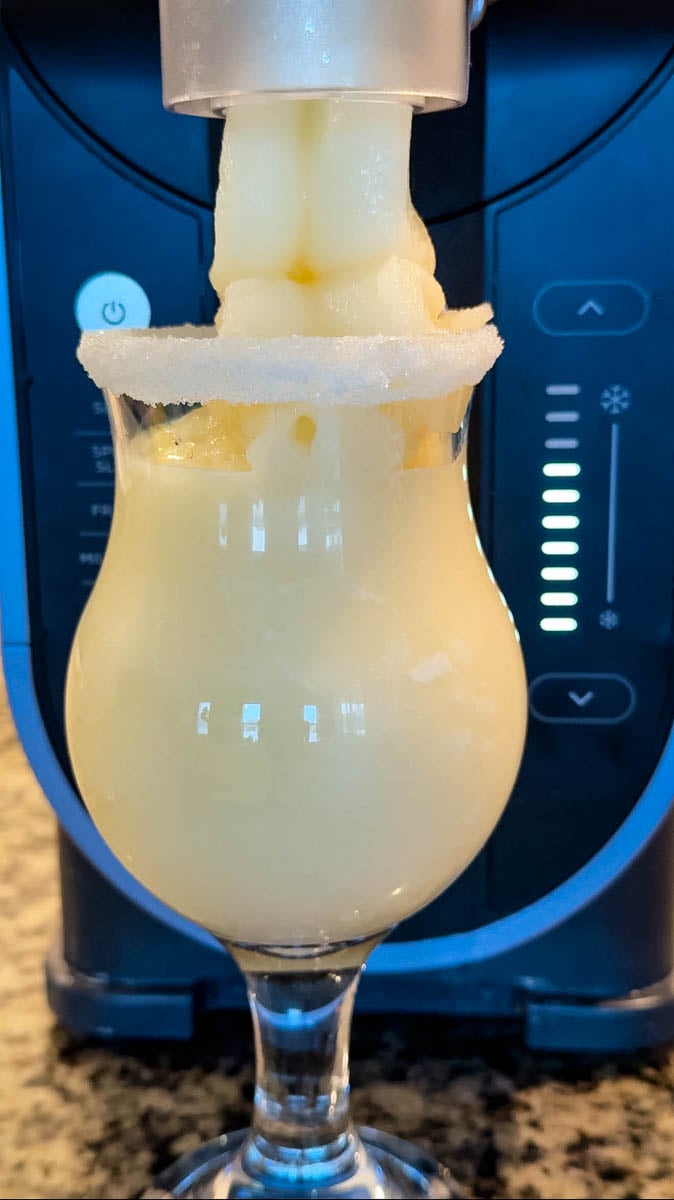 Piña colada slush dispensing from the Ninja Slushi machine into a sugar-rimmed glass.