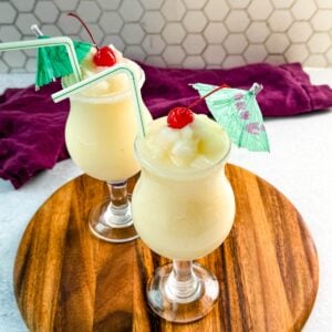 Close-up of creamy piña coladas in curved glasses with straws, cherries, and umbrellas, ready to serve.