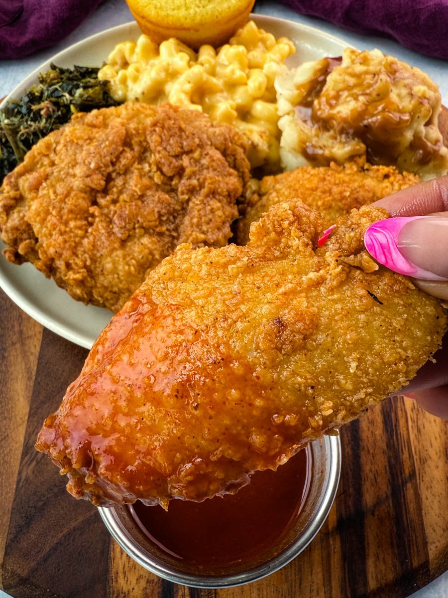 Close-up of a hand holding a freshly fried chicken wing dipped in hot sauce, with a plate in the background featuring fried chicken, mac and cheese, collard greens, mashed potatoes and gravy, and a cornbread muffin.