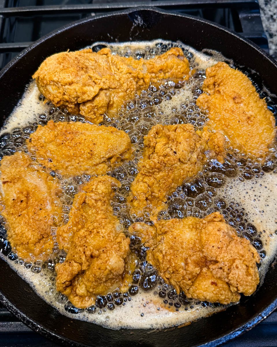Overhead shot of golden fried chicken pieces frying in hot oil in a cast iron skillet on the stovetop.
