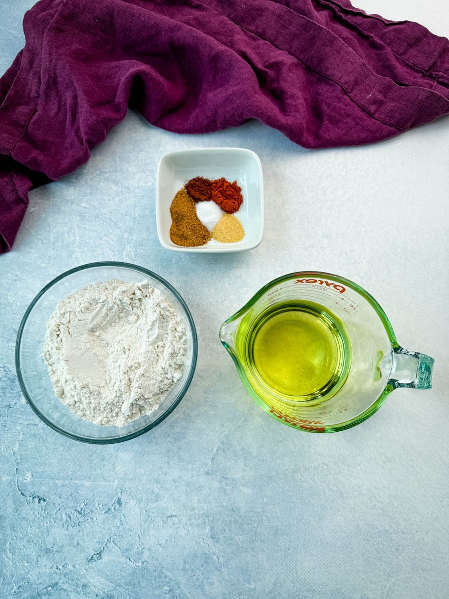 A bowl of all-purpose flour, a Pyrex measuring cup filled with cooking oil, and a dish of spices laid out on a counter next to a purple cloth—ingredients ready for coating and frying chicken.