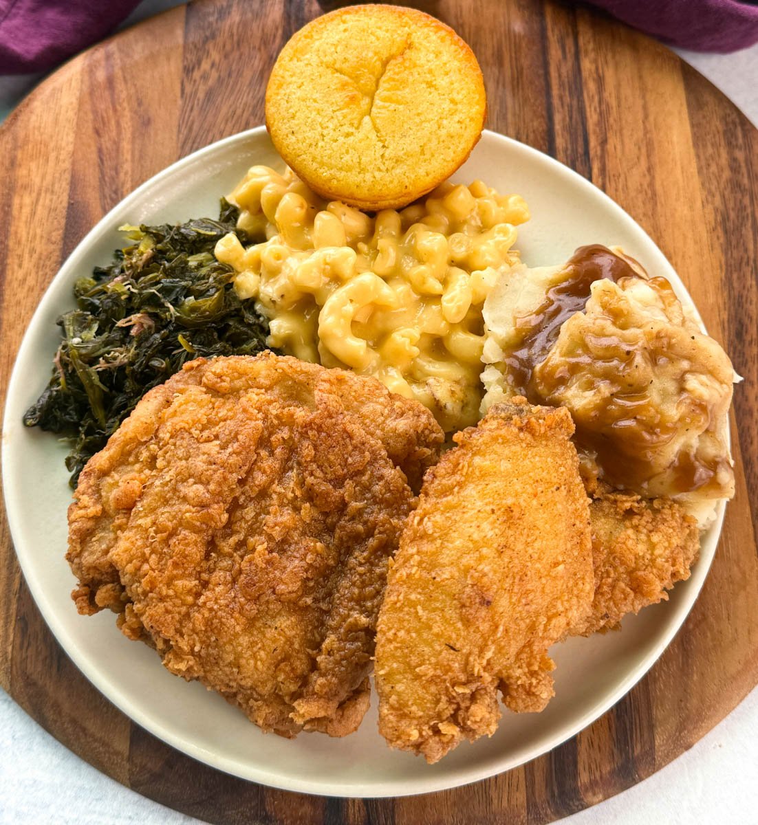 Overhead view of a full Southern dinner plate with golden fried chicken, creamy mac and cheese, collard greens, mashed potatoes with brown gravy, and a round cornbread muffin.