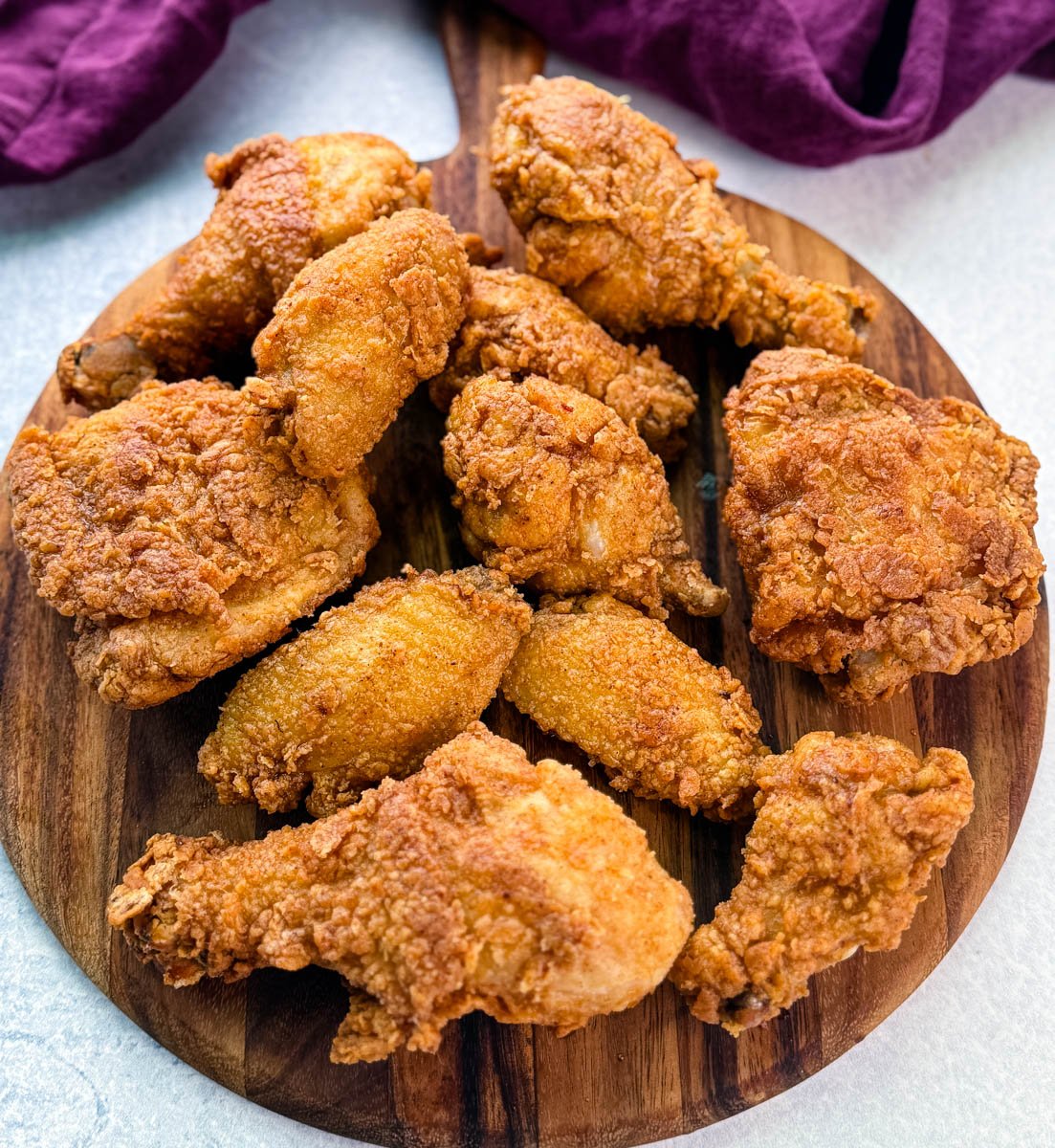 A wooden board topped with a spread of freshly fried, crispy chicken drumsticks, thighs, and wings, showcasing golden brown, crunchy coating.