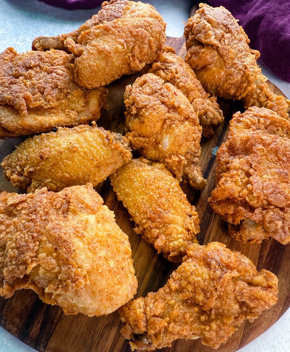Close-up view of several pieces of golden, crunchy fried chicken—wings, drumsticks, and thighs—arranged on a wooden serving board with a purple napkin in the background.