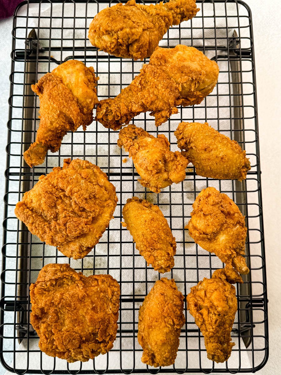 Multiple pieces of fried chicken spread across a wire rack for draining, showing their crispy, golden crust. Includes thighs, drumsticks, and wings.