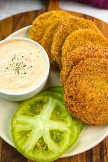 Well-styled plate of crispy fried green tomatoes next to a ramekin of seasoned dipping sauce and fresh tomato slices, ideal for serving at a Southern-style meal.