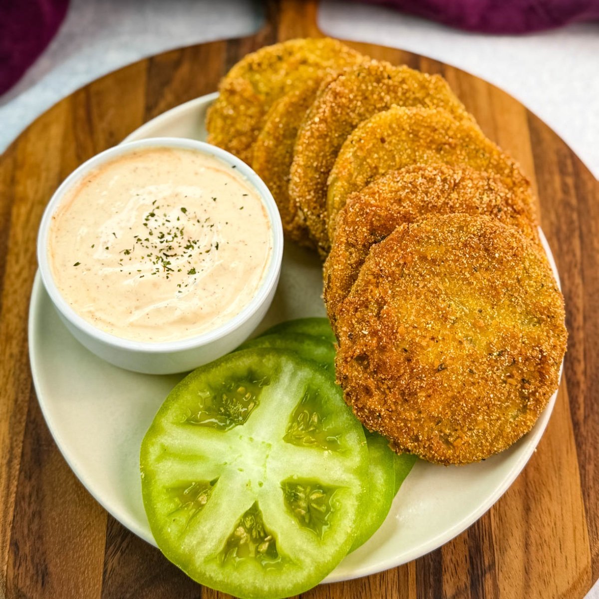 Well-styled plate of crispy fried green tomatoes next to a ramekin of seasoned dipping sauce and fresh tomato slices, ideal for serving at a Southern-style meal.