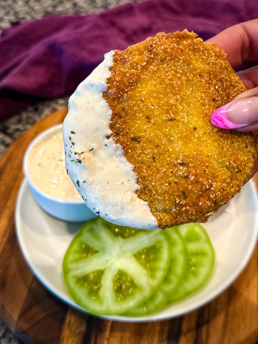 Close-up of a hand holding a crispy, golden-brown fried green tomato partially dipped in creamy white sauce, with sliced raw green tomatoes and dipping sauce in the background.