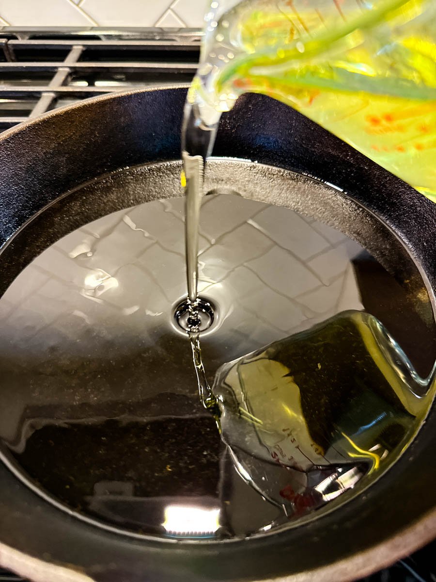 Action shot of canola oil being poured into a hot cast iron skillet, preparing for frying the tomatoes.
