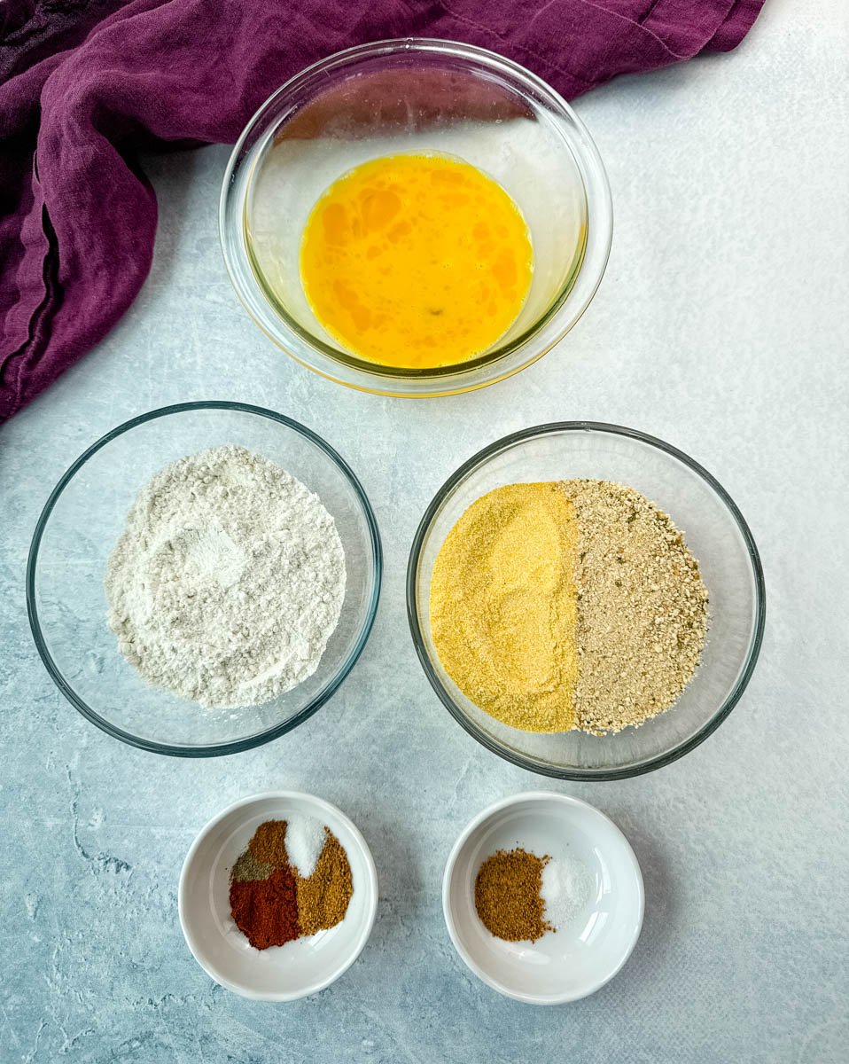 Overhead shot of breading station for fried green tomatoes featuring separate bowls with beaten eggs, all-purpose flour, a mix of yellow cornmeal and breadcrumbs, and small bowls of Creole seasoning and spices.