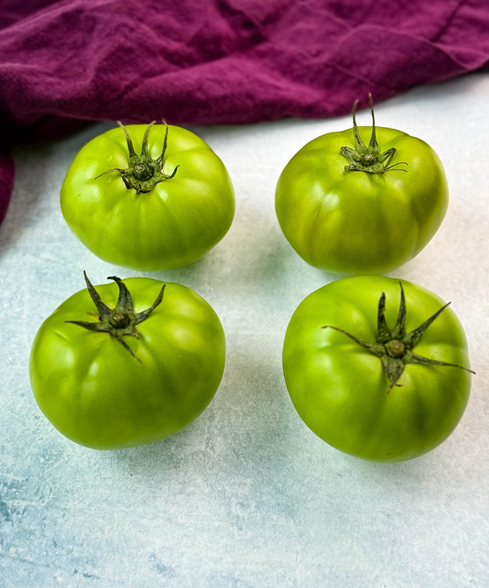 Four whole, unripe green tomatoes with stems intact placed on a white surface next to a purple kitchen towel—perfect for frying.