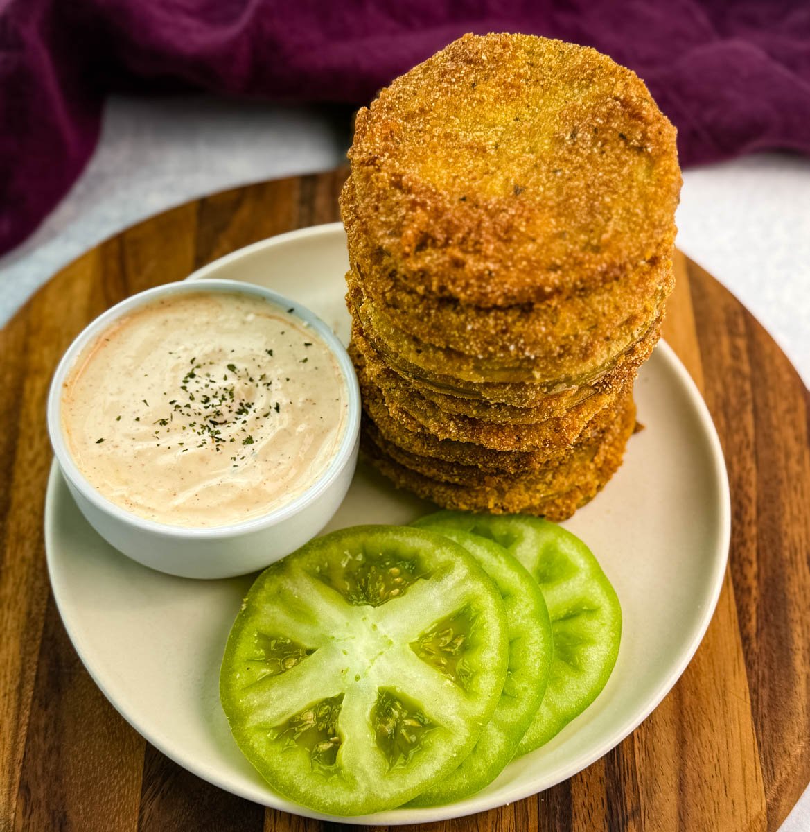 Tall stack of fried green tomatoes served on a white plate with creamy dipping sauce and fresh tomato slices, all on a wooden board with a purple kitchen towel nearby.