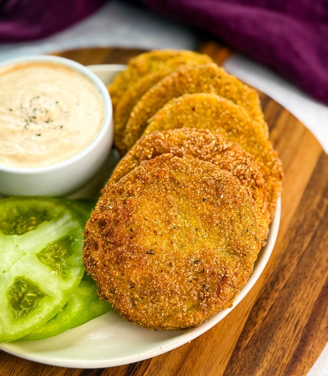 Golden fried green tomatoes arranged in a fanned-out line on a plate next to a bowl of dipping sauce and fresh green tomato slices.