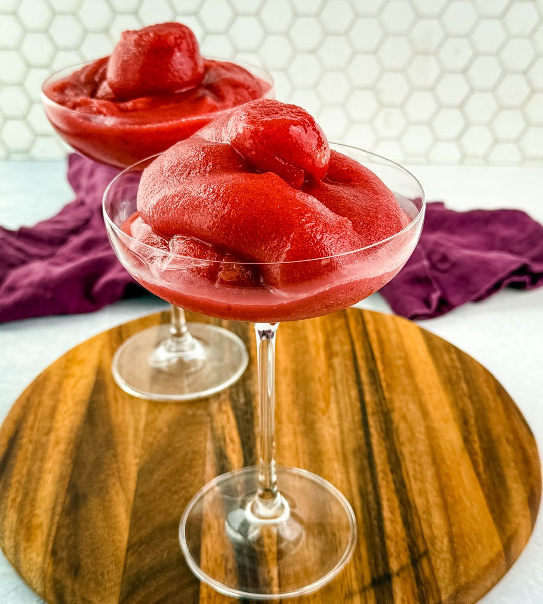 Two coupe glasses filled with thick, vibrant red frosé on a round wooden board with a purple cloth in the background.