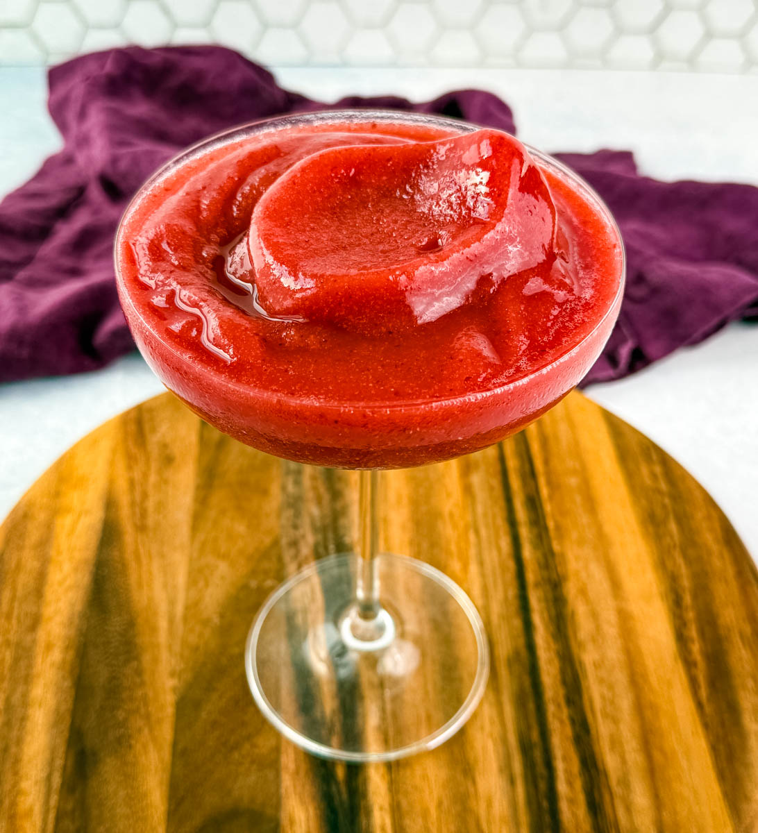 Single glass of thick, swirl-topped frosé on a wooden board with a hex tile background and a purple cloth draped nearby.