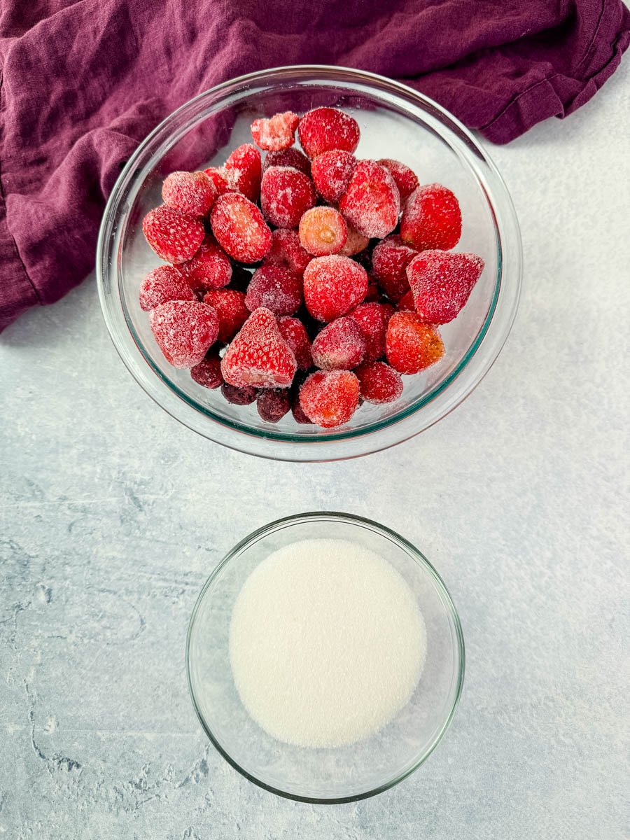 Overhead view of two glass bowls—one filled with frozen strawberries and the other with granulated sugar—set against a light background with a purple linen.