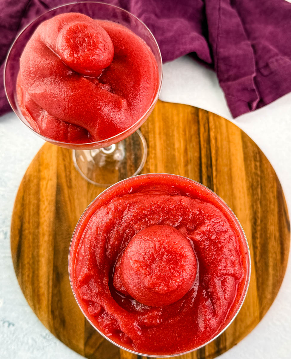Overhead view of two glasses filled with frosé, centered on a wooden board with visible texture and swirls in each glass.