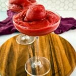 Front view of a single glass filled with a thick, icy red frosé, topped with a frozen berry swirl, another glass blurred in the background.