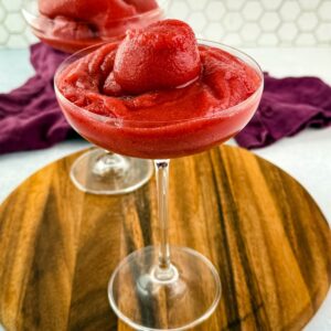 Front view of a single glass filled with a thick, icy red frosé, topped with a frozen berry swirl, another glass blurred in the background.