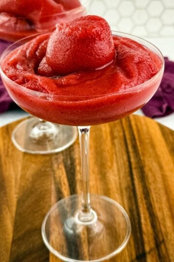 Front view of a single glass filled with a thick, icy red frosé, topped with a frozen berry swirl, another glass blurred in the background.