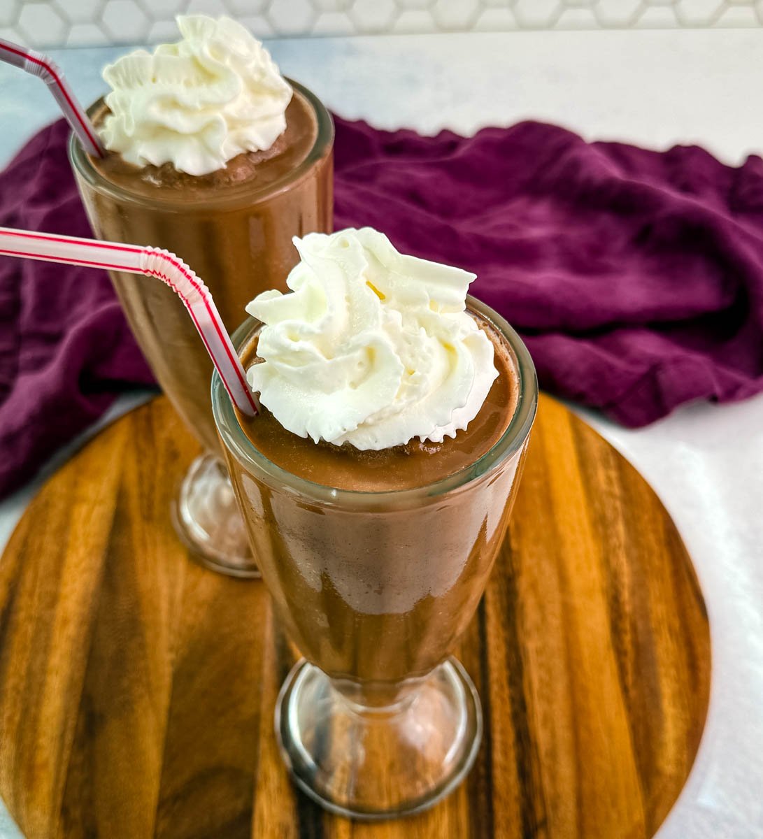 Top-down view of two chocolate milkshakes with whipped cream and red-striped straws, sitting on a round wooden board with a purple napkin in the background.
