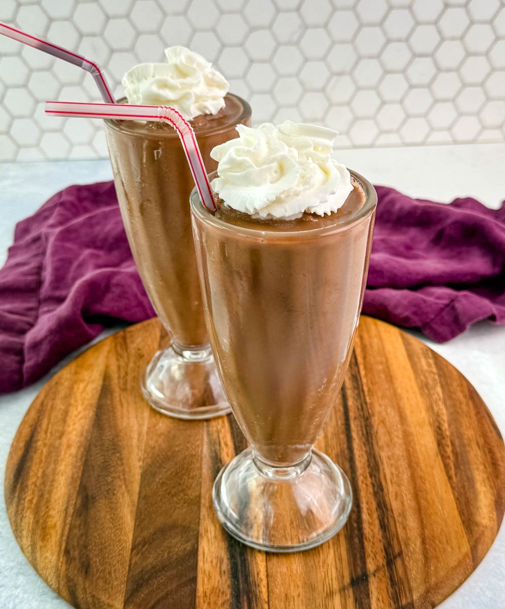 Two tall glass milkshake glasses filled with rich chocolate milkshake, topped with whipped cream and red-striped straws, set on a wooden tray with a purple cloth in the background.