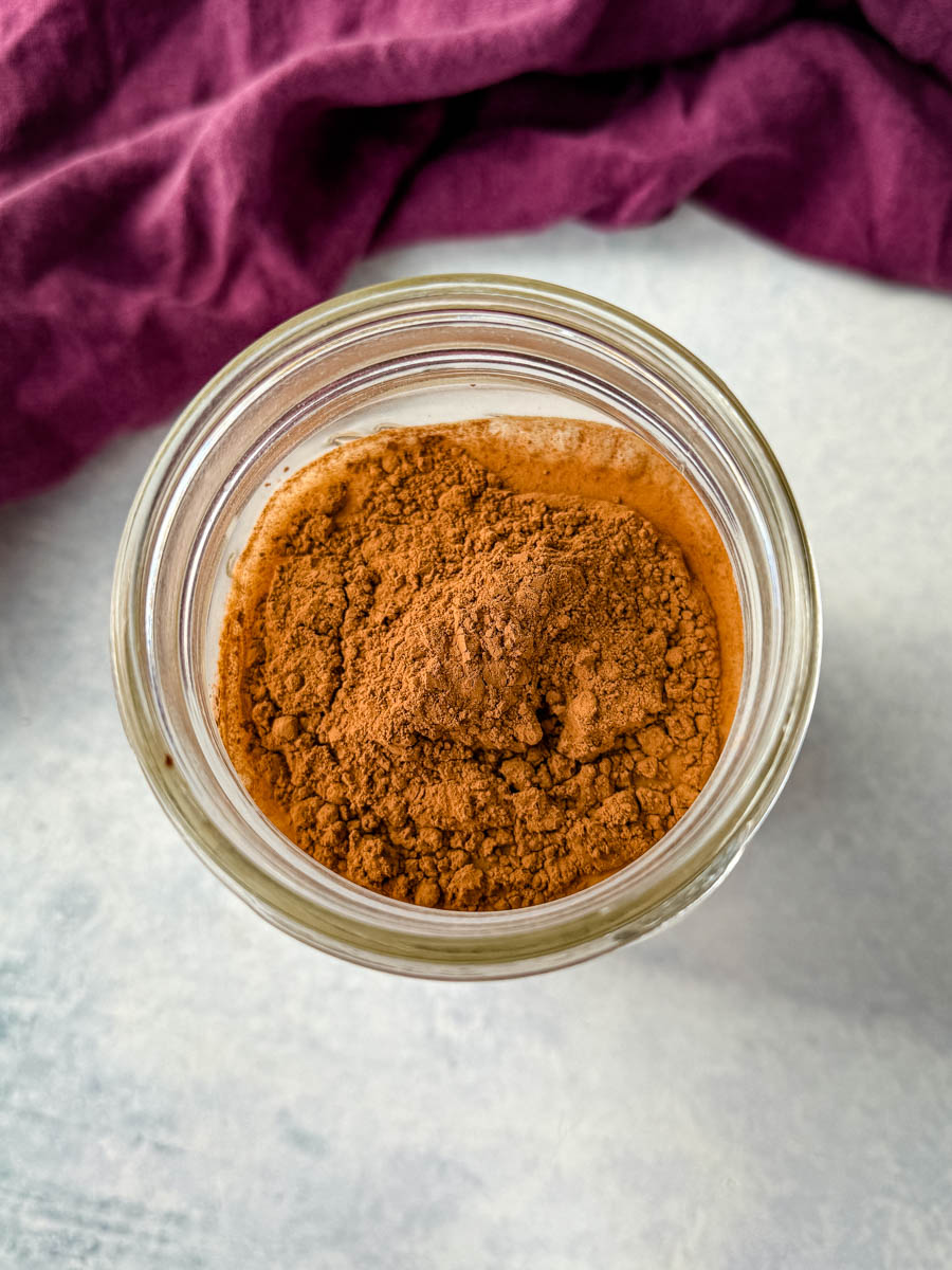 Overhead view of a small glass jar filled with unsweetened cocoa powder, set against a light surface with a deep purple cloth in the background.
