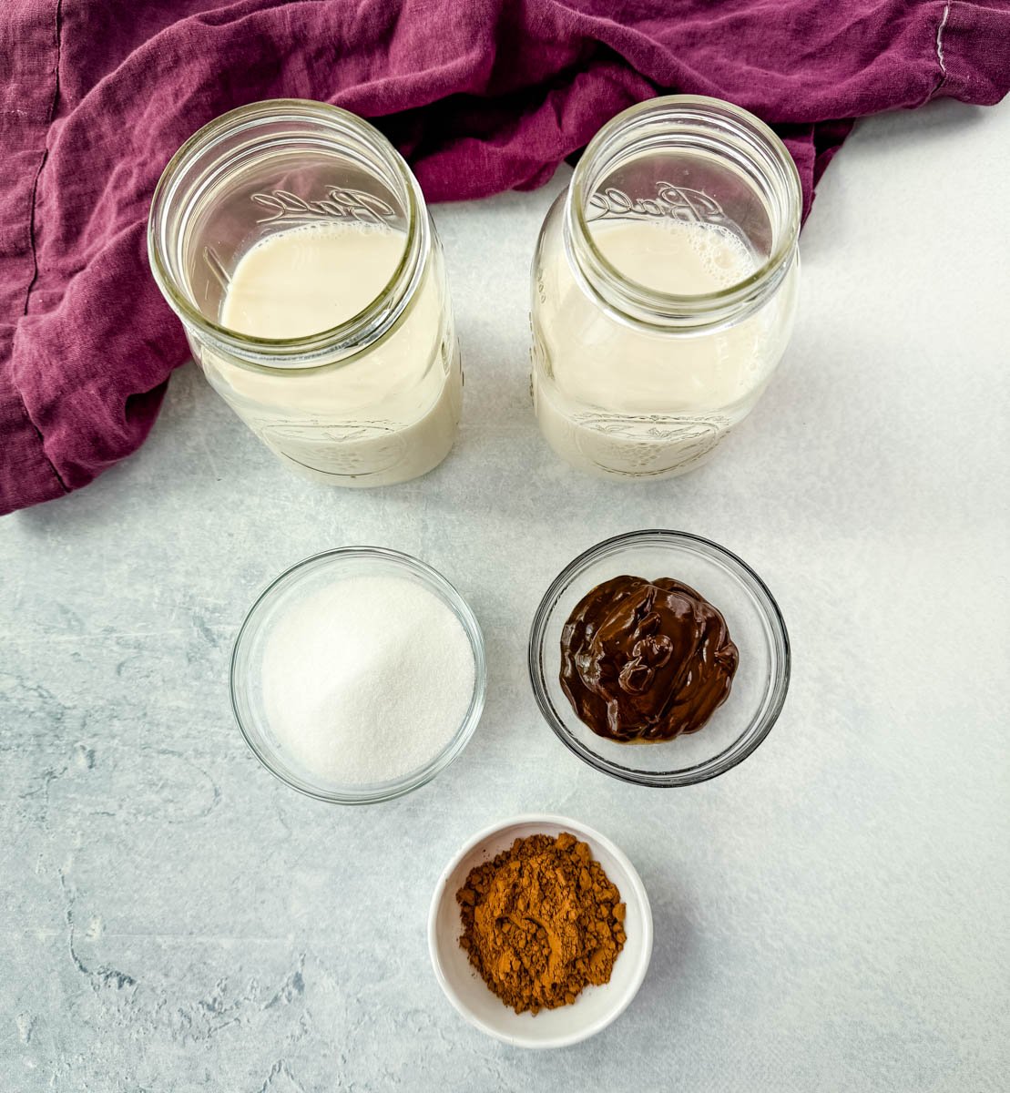 Overhead layout of chocolate milkshake ingredients including two mason jars filled with milk, a small bowl of sugar, a bowl of chocolate syrup, and a dish of cocoa powder, all placed on a light surface with a purple cloth nearby.