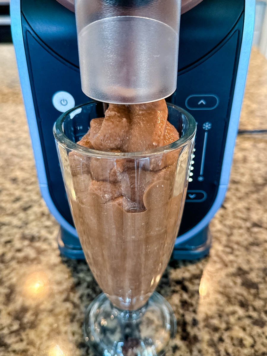 A chocolate milkshake being dispensed directly into a glass from the Ninja Slushi Machine, showcasing the smooth frozen texture and machine design.