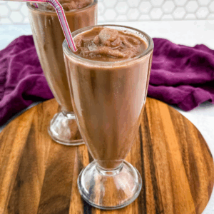 Two chocolate milkshakes served in classic soda fountain glasses on a wood platter, showing off the thick, frozen texture and striped straws.