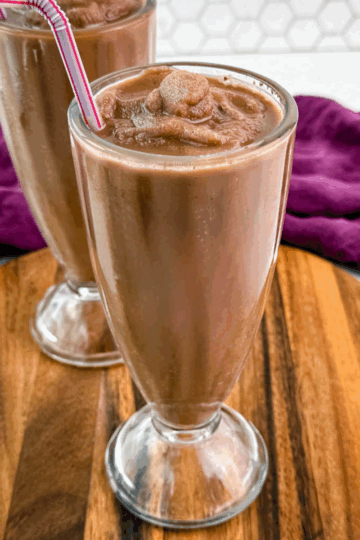 Two chocolate milkshakes served in classic soda fountain glasses on a wood platter, showing off the thick, frozen texture and striped straws.