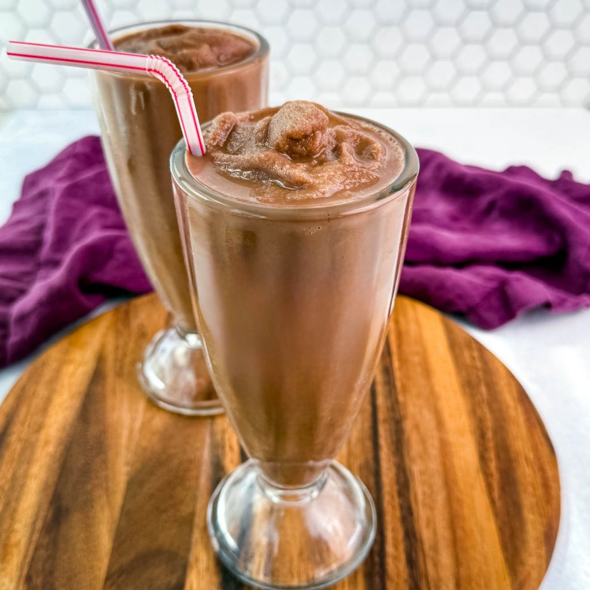 Two chocolate milkshakes served in classic soda fountain glasses on a wood platter, showing off the thick, frozen texture and striped straws.