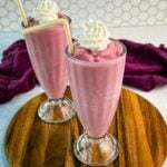 Two tall milkshake glasses filled with thick pink strawberry milkshakes, topped with whipped cream and yellow-striped straws, placed on a round wooden board with a purple cloth in the background.
