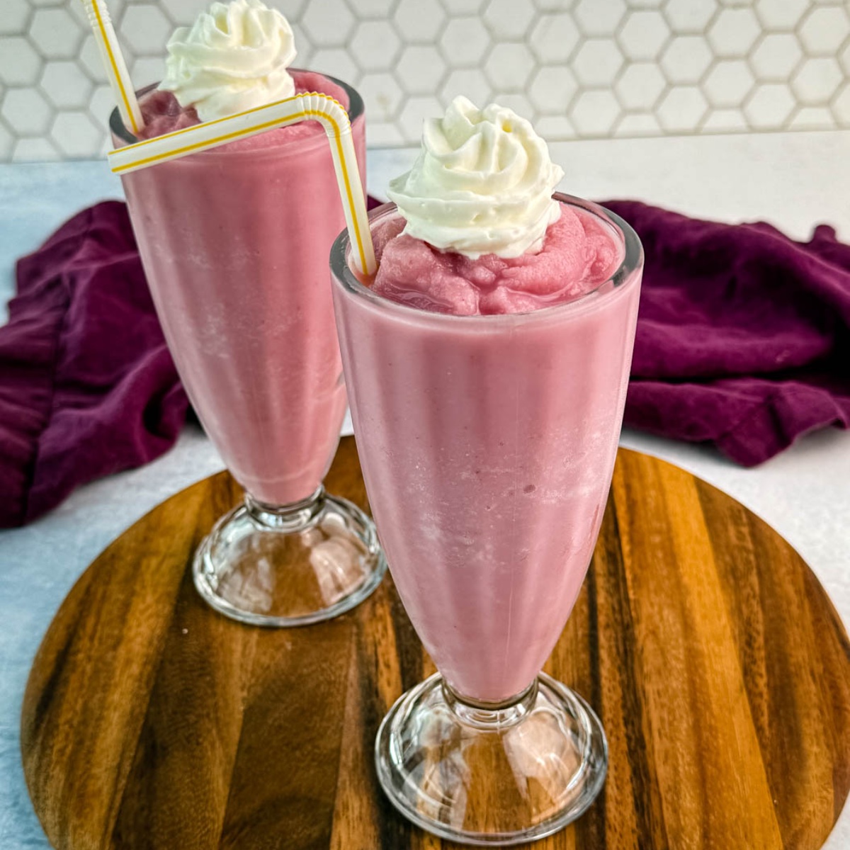 Two tall milkshake glasses filled with thick pink strawberry milkshakes, topped with whipped cream and yellow-striped straws, placed on a round wooden board with a purple cloth in the background.