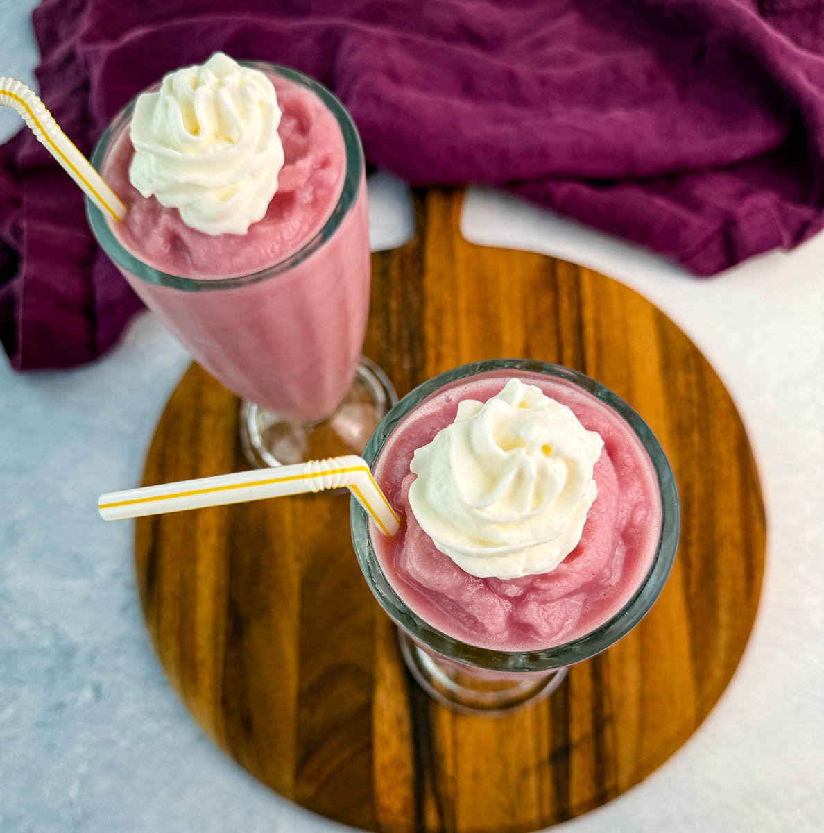 Overhead view of two pink milkshakes topped with whipped cream and straws, sitting on a wooden board with a purple cloth to the side.