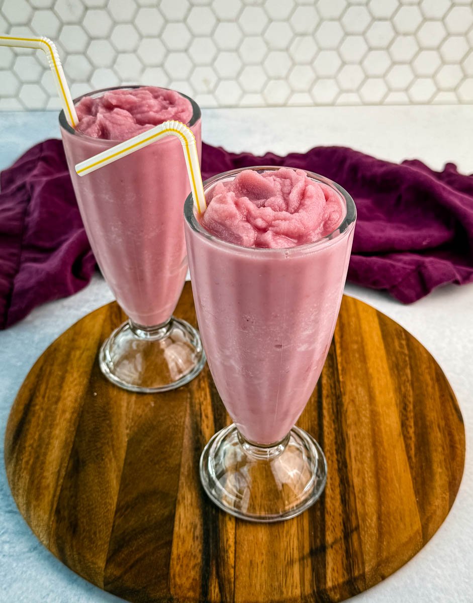 Two tall glasses filled with thick, slushy pink strawberry milkshakes without whipped cream, placed on a round wooden board with a hexagonal tile background.
