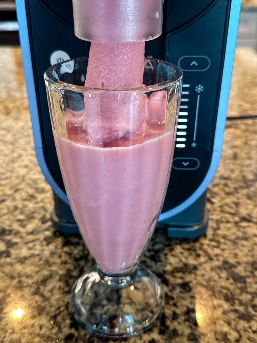 A tall glass being filled with a frozen strawberry milkshake mixture directly from the Ninja Slushi machine’s spout, showing the soft, frozen texture as it pours in.