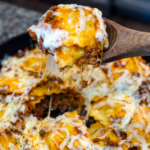 Close-up of a cheesy beef and ravioli lasagna being scooped from a cast iron skillet with a wooden spoon. Melted cheese is stretched with bits of seasoned ground beef
