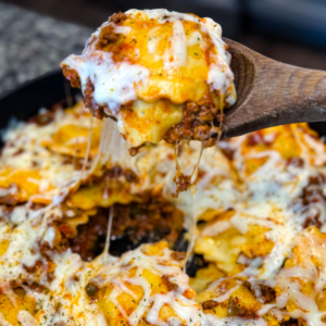 Close-up of a cheesy beef and ravioli lasagna being scooped from a cast iron skillet with a wooden spoon. Melted cheese is stretched with bits of seasoned ground beef