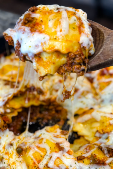 Close-up of a cheesy beef and ravioli lasagna being scooped from a cast iron skillet with a wooden spoon. Melted cheese is stretched with bits of seasoned ground beef