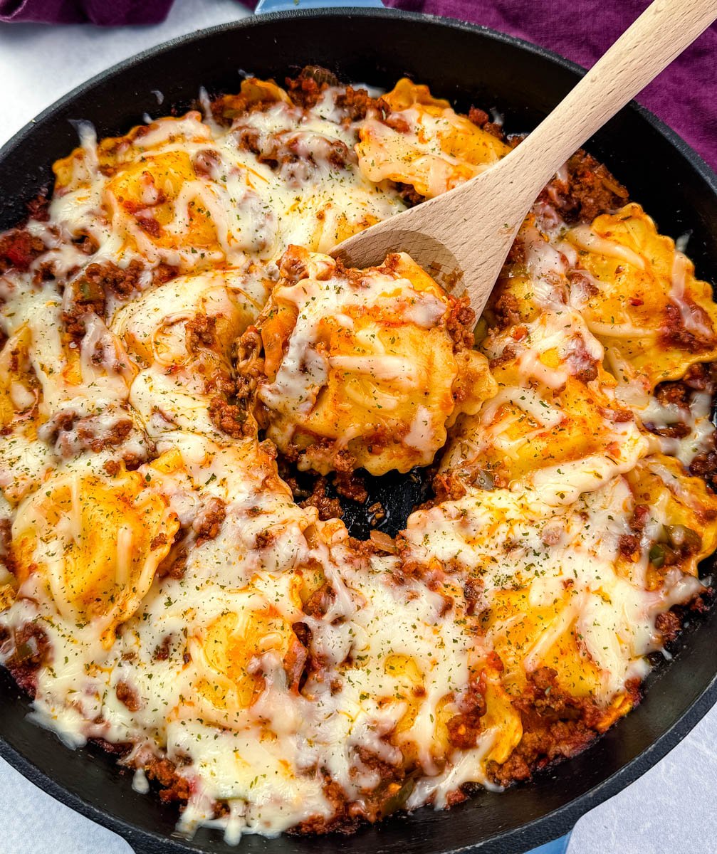 Overhead view of one-pot ravioli lasagna in a skillet, covered in melted cheese and herbs. A wooden spoon is lifting a portion from the center.
