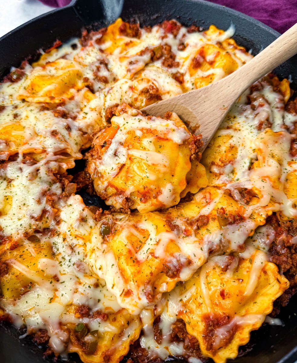 Top-down view of baked ravioli lasagna in a skillet. Ravioli is layered with meat sauce and melted cheese, garnished with dried herbs.