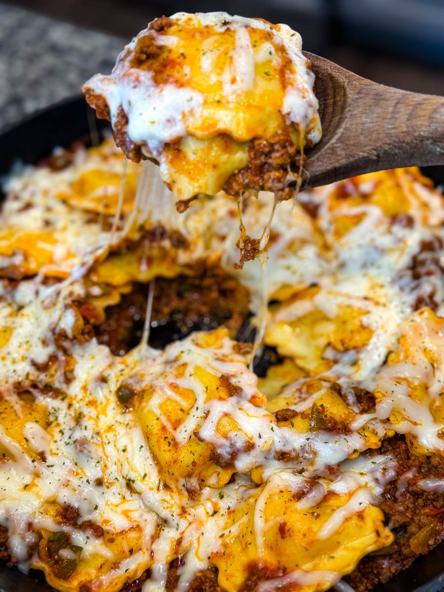 Dramatic close-up of a gooey, cheesy scoop of ravioli lasagna being lifted from the skillet, with melted cheese stretching from the pan.
