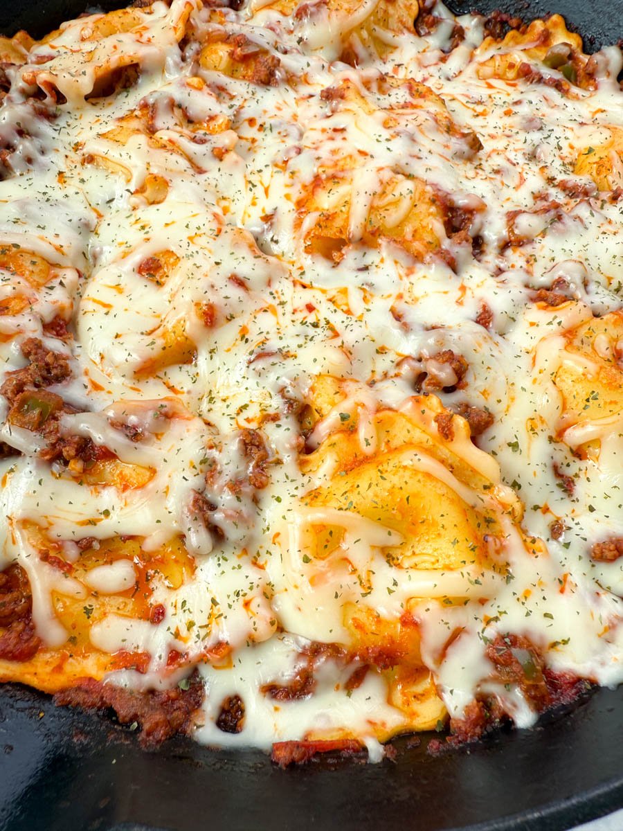 Overhead view of a finished one-pot ravioli lasagna in a cast iron skillet, topped with melted mozzarella cheese and garnished with herbs.
