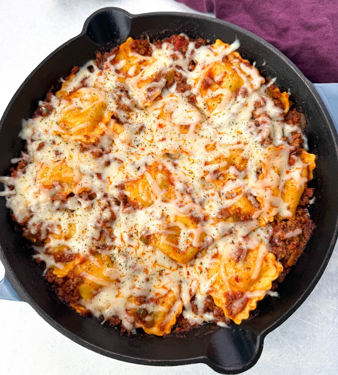 Overhead view of a finished one-pot ravioli lasagna in a cast iron skillet, topped with melted mozzarella cheese and garnished with herbs.