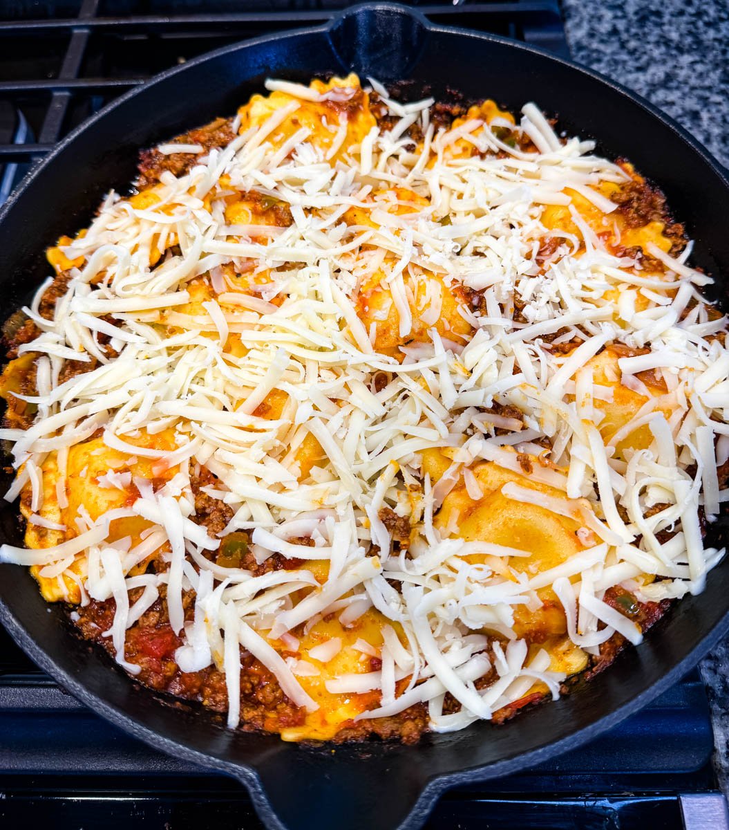 Unbaked ravioli lasagna in a cast iron skillet with shredded mozzarella cheese layered over the top, ready to be cooked.